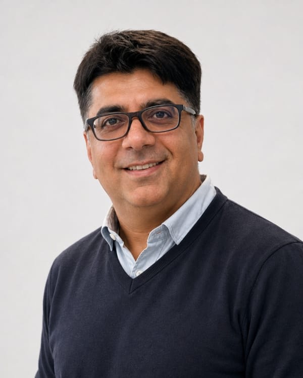 Kamal Hans smiling, wearing glasses and a dark sweater over a light collared shirt